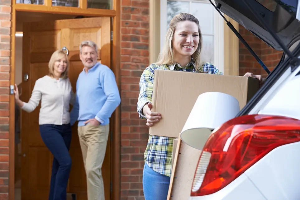 Young woman loads moving box into car while parents look on