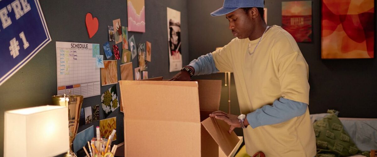 A college student in his decorated dorm organizing a cardboard box on top of his desk.