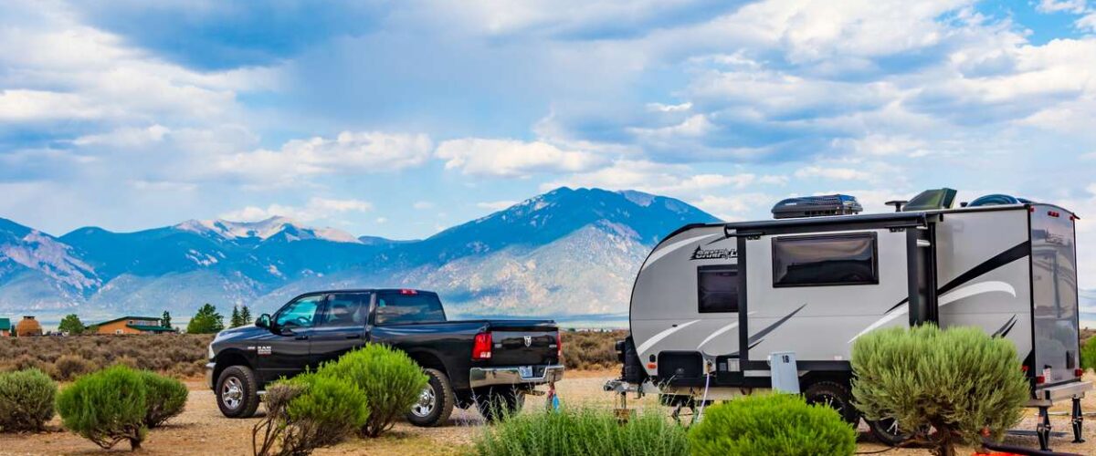 A blue pickup truck and an RV are parked in a desert on a bright day with mountains behind.