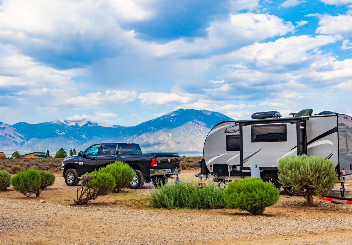 A blue pickup truck and an RV are parked in a desert on a bright day with mountains behind.