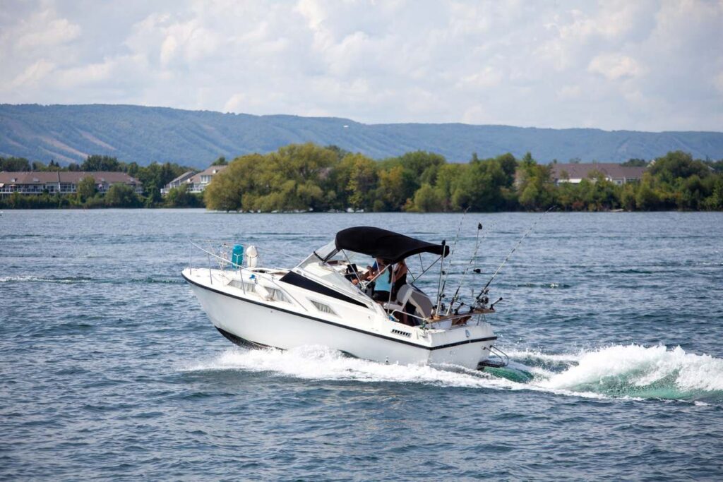 A small white speed boat glides across the water of a lake with trees lining the water and mountains in the distance.
